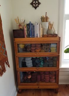 a wooden shelf with yarn and books on it next to a window in a room