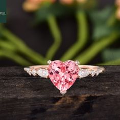 a pink heart shaped diamond ring on top of a piece of wood with flowers in the background