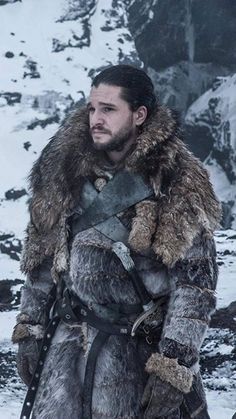 a man dressed in fur and leather standing on snow covered ground with mountains behind him