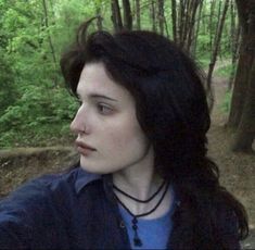 a woman with long black hair standing in the woods looking off into the distance while wearing a blue shirt