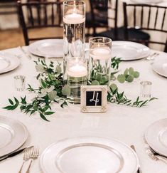 the table is set with white plates and silverware, candles and greenery on them