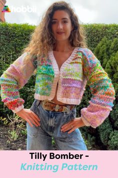 a woman standing in front of bushes wearing a colorful knitted sweater and jean shorts