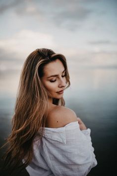 a woman with long hair standing in front of the ocean looking off into the distance