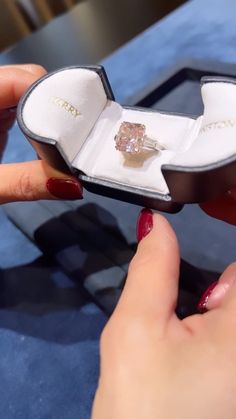 a woman's hand holding an open ring box with a pink diamond in it