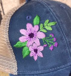 Hand embroidered  Charming purple flowers are embroidered on the front of this mesh-back ponytail hat. Hook and loop adjustable back. Women's fit Washed cotton twill front panels Soft mesh back panels Ponytail opening Structured, mid to low-profile, six-panel Pre-curved visor Micro hook and loop closure Spring Cotton Snapback Trucker Hat, Cotton Snapback Trucker Hat For Spring, Spring Cotton Trucker Hat With Curved Brim, Embroidered Adjustable Trucker Hat, Summer Purple Baseball Cap, Embroidered Summer Trucker Hat With Curved Bill, Custom Embroidered Summer Hats With Adjustable Fit, Summer Embroidered Trucker Hat, Custom Embroidery Summer Baseball Cap