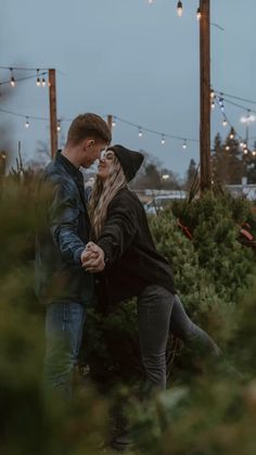 a young man and woman embracing in front of christmas trees at an outdoor market with lights strung overhead