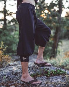 a man standing on top of a rock in the woods with his legs crossed and wearing flip flops