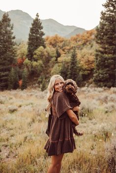 a woman holding a dog in her arms while standing in a field with mountains behind her