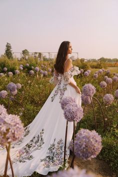 a woman is standing in a field with purple flowers and wearing a white dress that has floral embroidery on it