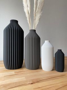 three vases are lined up on a table with long grass in them and one is black, the other white