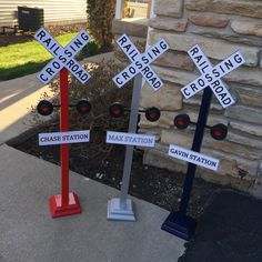 three railroad crossing signs in front of a brick building with grass and bushes behind them