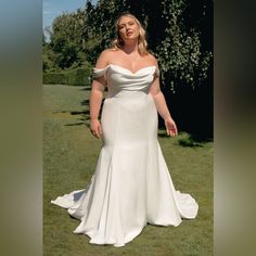 a woman in a white wedding dress standing on the grass with her hands behind her back