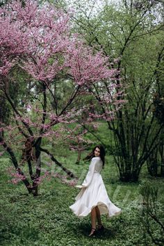 a woman in a white dress is walking through the grass and trees with pink flowers