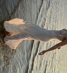 a woman in white dress walking on beach next to the ocean with her legs spread out