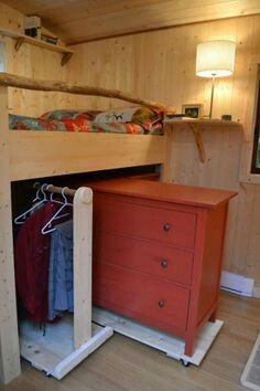 a red dresser sitting under a bed next to a wooden shelf with clothes on it