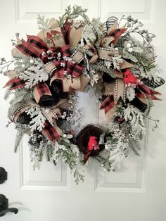 a christmas wreath hanging on the front door with red and white ribbon, evergreens, pine cones and bells