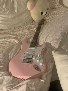 a pink guitar sitting on top of a bed next to a teddy bear toy and pillow