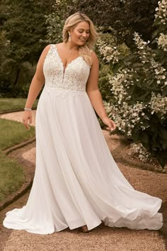 a woman in a white dress standing on a path with trees and bushes behind her