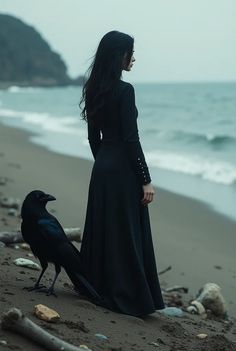 a woman standing on the beach next to a black bird and looking at the ocean