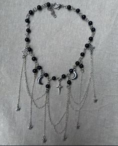 The necklace has a celestial-inspired design, featuring black beads connected by silver chain links. It includes various hanging silver charms, such as crescent moons, stars, and a central starburst or compass-shaped charm. Several delicate silver chains drape down in layers, adding an elegant, flowing effect to the piece. The overall look is both mystical and stylish, blending dark tones with metallic details for a balanced, striking appearance. Goth Necklace, Silver Chains, Chain Links, Dream Jewelry, Black Beads, Silver Charms, Romania, Chain Link, Silver Chain