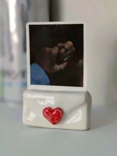 a small white photo frame with a red heart on the front, and an image of a person holding a baby's hand