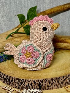 a small crocheted chicken sitting on top of a tree stump next to flowers