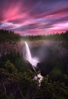 a waterfall in the middle of a forest under a purple and pink sky with clouds