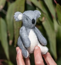 a person holding a small stuffed koala bear in their left hand with green leaves behind it