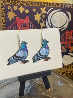 two blue birds with chains hanging from their earring hooks on a white card in front of a painting