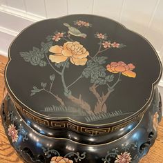 an ornate black box with flowers painted on the lid sits on a wooden floor next to a white wall