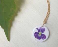 "This is my original design of a tiny, sweet violet flower. I am fascinated by violets; their deep jewel-like intense colors, delicate petals, lush heart-shaped leaves, and spindly stems. A big patch of violets comes up in our backyard every spring and when they bloom, my older daughter picks a handful for me to put in my small glass vase on the windowsill by the kitchen sink. They are the brightest color in the room! This is one of my smallest pendants; it measures 5/8\" across, smaller than a Purple Flower Necklace With Flower Charm, Purple Flower Charm Necklace, Purple Flower Pendant Necklace, Purple Flower Charm Necklace As A Gift, Handmade Purple Flower Pendant Necklace, Purple Flower Pendant With Pressed Flowers, Purple Flower Shaped Necklace With Pressed Flowers, Handmade Delicate Purple Necklace, Purple Flower Pendant Necklace For Birth Flower