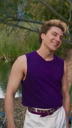 a young man standing in front of a body of water wearing a purple shirt and white shorts