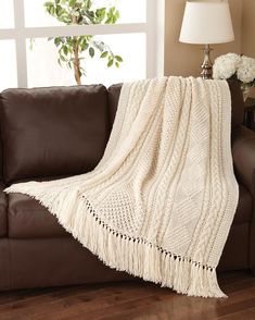 a white blanket sitting on top of a brown couch next to a vase with flowers