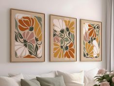 three floral paintings hang on the wall above a white couch in front of a window