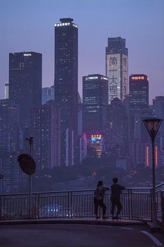 two people are standing on a bridge looking at the city lights and skyscrapers in the distance