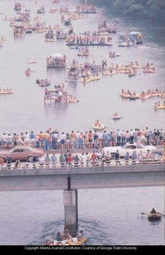 a large group of people standing on the side of a bridge over water with boats in it