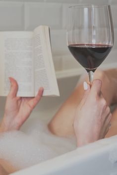 a woman laying in a bathtub reading a book and holding a glass of wine