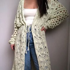 a woman standing in front of a wall wearing a long cardigan sweater and jeans