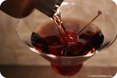 someone is pouring liquid into a martini glass with cherries on the rim and a spatula in it