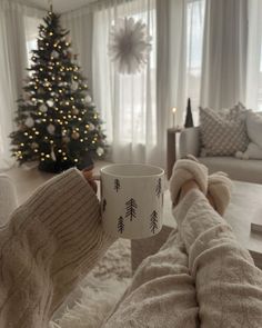 a person holding a coffee cup in their hand while sitting on a couch with a christmas tree behind them