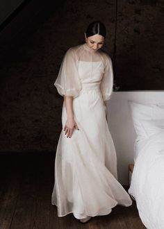 a woman in a white dress standing next to a bed and looking down at the floor