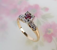 "A sparkling red spinel gemstone is centered on this vintage 1940s 14k yellow/white gold diamond ring. The Heintz Bros. Inc. (est. 1934, Buffalo, NY) made the vintage setting with a raised pierced solitaire gallery and classic styling. The .10 carat natural spinel gem is of deep pink to red color and measures approx. 2.7mm round with a 1.8mm depth. Each shoulder is set with one .01 carat 1.8mm diamond. The gemstone settings are of white gold and the sturdy band is of yellow gold. The ring measur Vintage Ruby Ring With Diamond Accents, Vintage Ruby Ring With Diamond Accents For Wedding, Vintage 14k Gold Ruby Ring With Accent Stones, Classic Red Birthstone Ring With Diamond Accents, Heirloom Style Red Diamond Ring With Accents, Heirloom Red Diamond Ring With Accents, Vintage Ruby Rings With Diamond Accents, Vintage Ruby Ring With Accent Stones For Anniversary, Vintage Red Ruby Ring For Anniversary