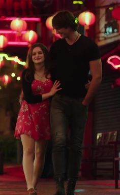 a young man and woman walking down the street at night with red lights in the background