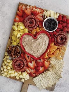 a heart shaped platter filled with fruit, crackers and dips on a cutting board