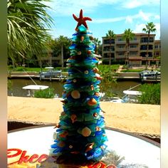 a christmas tree made out of plastic bottles on a table near the water with boats in the background