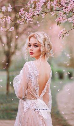 a woman in a white dress standing under a tree with pink flowers on it's branches