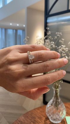 a woman's hand with a diamond ring on top of her finger, in front of a vase filled with flowers