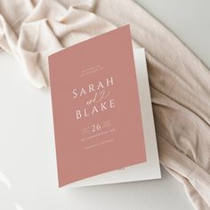 a pink and white wedding card is laying on top of a bed next to a blanket