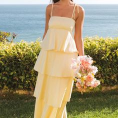 a woman in a yellow dress standing on grass near the ocean holding a bouquet of flowers