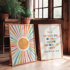 two framed art prints on the floor in front of a potted plant and window